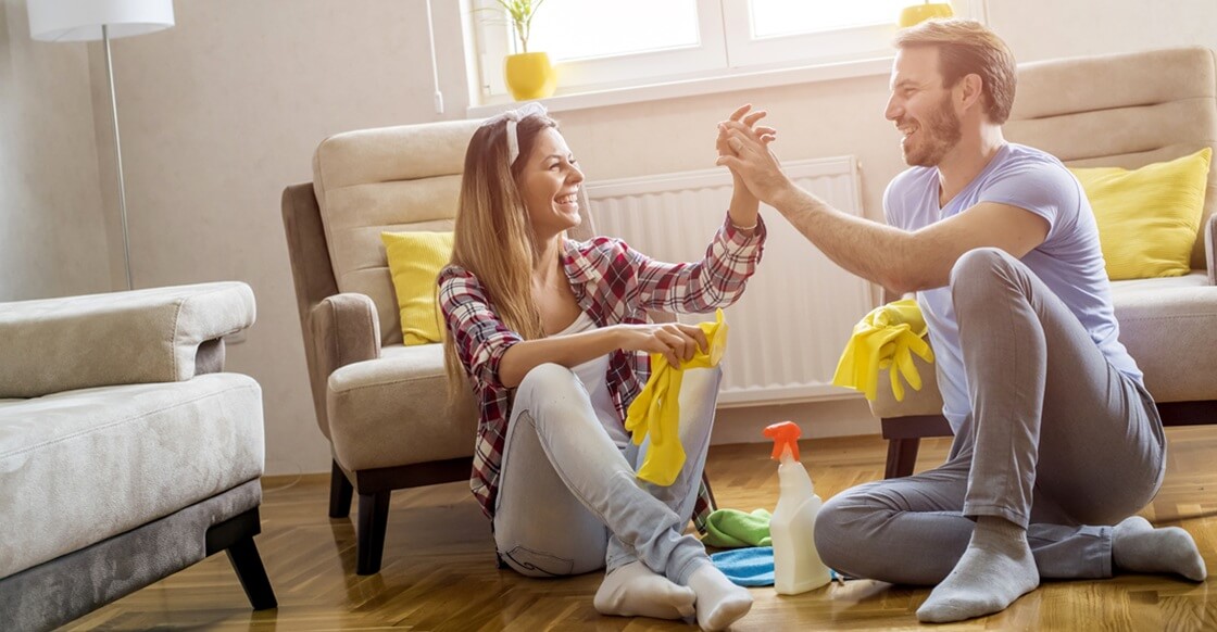 Smiling couple sitting on the floor of their living room during spring cleaning, surrounded by cleaning supplies, sharing a high-five after tidying up.